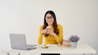 Çekici ve mutlu Asyalı genç bir bayan üniversite öğrencisi ya da gözlüklü bir çalışan masasında otururken kahve fincanını tutuyor. beyaz arkaplanda izole edilmiş