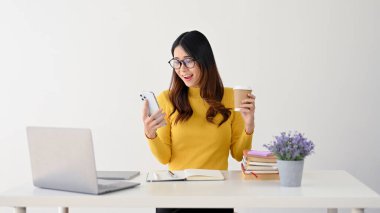 Neşeli ve heyecanlı bir Asyalı genç bayan öğrenci ya da çalışan masasında kahvesini yudumlarken telefonundaki inanılmaz ve beklenmedik bir mesaja bakıyor. beyaz arkaplanda izole edilmiş