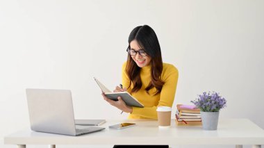 Gözlüklü güzel ve neşeli bir Asyalı kız üniversite öğrencisi masasında otururken kitabına notlar alıyor. beyaz arkaplanda izole edilmiş