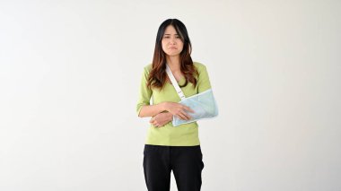 Kol askısında kırık koluyla sıradan kıyafetler giyen üzgün ve incinmiş genç bir Asyalı kadın izole edilmiş beyaz bir arka plana karşı duruyor. kol yaralanması, kol ağrısı, sağlık sigortası, kaza