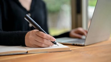 Bir iş kadınının kalem tutarken ve görevlerini not defterine yazarken yakın plan görüntüsü. Masasındaki dizüstü bilgisayarında çalışırken..