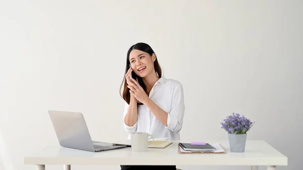 An attractive Asian businesswoman or female assistant in a white shirt is talking on the phone with a client at her desk. isolated white studio background