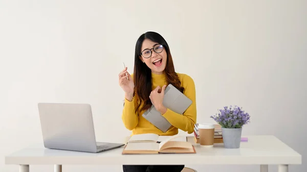 stock image A cheerful and charming young Asian female in glasses and casual clothes sits at her working desk with her laptop, tablet, books and stuff. college student, freelancer, businesswoman