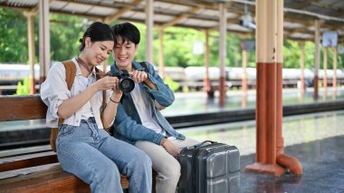 Güzel, genç bir Asyalı çift tren istasyonunda trenlerini beklerken kamerada konuşuyor ve resimlerine bakıyor. Sırt çantası, Gezginler, Turist, Tatil, Tatil
