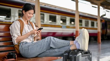Güzel ve mutlu bir Asyalı genç bayan sırt çantalı ya da gezgin akıllı telefonunu kullanıyor ya da trenini platformda beklerken seyahat rehberi web sitesine kayıyor..