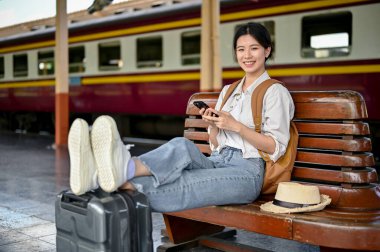 Güzel ve mutlu bir Asyalı genç bayan gezgin akıllı telefonunu bir bankta dinlenirken tren istasyonunda trenini beklerken kullanıyor..