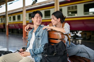 Genç ve sevimli bir Asyalı turist çifti trene birlikte binmeden önce platformda bir bankta konuşmaktan zevk alıyor. Balayı gezisi, turist çifti, seyahat partneri, ya da arkadaşlar, erkek arkadaş ve kız arkadaş