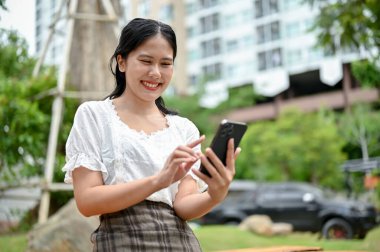 Güzel ve mutlu bir Asyalı kadının portresi. Günlük giysiler içinde. Akıllı telefonunu açık hava bahçesinde dinlenirken kullanıyor..