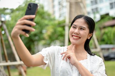 Güzel ve neşeli bir Asyalı kadın, günlük kıyafetleriyle resim çekiyor ya da şehir parkında dinlenirken akıllı telefonuyla fotoğraf çekiyor..