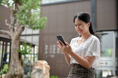 Mutlu bir Asyalı kadın telefon ekranına bakıyor, dışarıda dinlenirken mesajlar okuyor ya da biriyle sohbet ediyor..