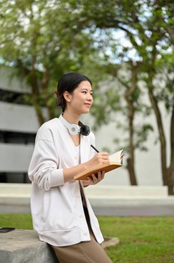 Kampüs parkındaki bir masada elinde not defteriyle oturan güzel ve mutlu bir Asyalı üniversite öğrencisi portresi..