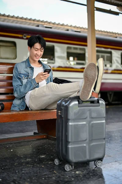 A handsome and happy young Asian male backpacker or traveler using his smartphone or scrolling on a travel guide website while waiting for his train at the platform. Solo travel, Summer vacation