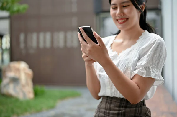 一个穿着休闲装的快乐的年轻亚洲女人用她的智能手机 一边和她的朋友聊天一边在公园里放松 — 图库照片