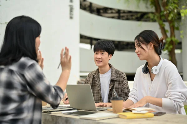 stock image A cheerful and positive young Asian male college student is listening to his friend's opinion while working on their group project at a table in the campus park together.