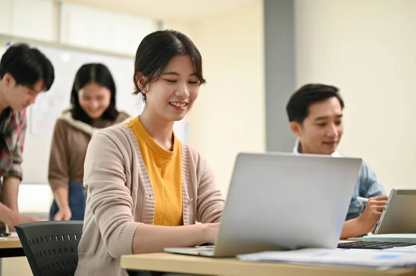 一位年轻貌美 聪明聪明的亚洲女员工在一家初创公司工作时 同事们还在房间里 她正在用笔记本电脑完成自己的任务 — 图库照片