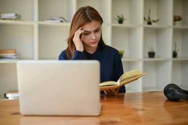 Stresli ve ciddi bir Asyalı kız üniversite öğrencisi kütüphanedeki masasında kitap okumaya odaklanıyor. eğitim kavramı