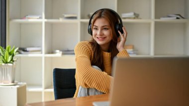 Mutlu ve güzel sarı kazaklı Asyalı bir kadın evde bir masada otururken kulaklıkla müzik dinlemekten zevk alıyor..