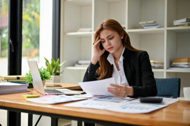Asyalı stresli ve yorgun bir iş kadını ofisindeki masasında mali raporları okumaya ve analiz etmeye odaklanıyor..
