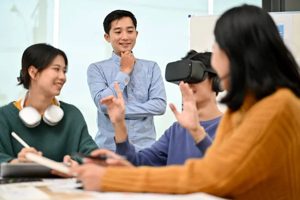 A cheerful Asian male developer sitting at the table tested a new VR game with virtual reality goggles in the meeting with his team. Virtual reality innovation, tech gadget