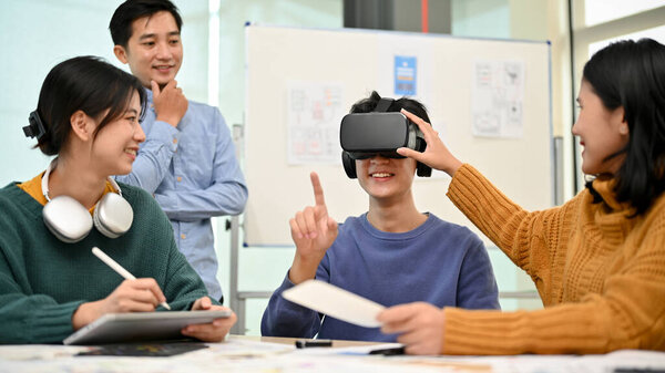 A team of Asian developers is in the meeting and excited about their newly developed VR game while a male colleague testes it with virtual reality goggles.