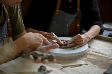 Yakın çekim görüntüsü, küçük bir çömlek çarkına işlenmemiş kil şekillendiren ya da yontan bir adam, arkadaşıyla birlikte bir atölyede el yapımı kil süsü yapıyor..