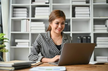 Kendine güvenen ve profesyonel Asyalı bir iş kadını ya da kadın patron e-postalarına cevap veriyor ve modern ofis odasındaki bilgisayarını kullanıyor..