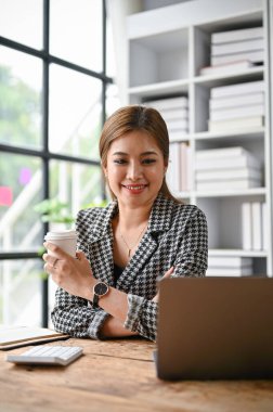 Güzel ve profesyonel bir Asyalı iş kadınının portresi. Masasındaki internet sitesinde iş makalesini okurken kahvesini yudumluyor..
