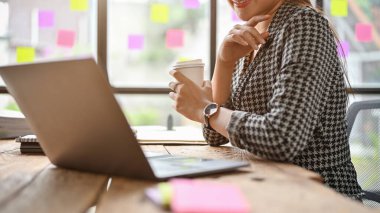 Asyalı profesyonel bir iş kadınının, kahve yudumlarken ve ofisteki bilgisayarında e-postalarını kontrol ederken çekilmiş yan görüntüsü..