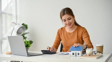 Masasında çalışan güzel ve mutlu Asyalı bir emlakçı kadın peşinatı hesaplıyor ve müşterileri için garanti belgesi hazırlıyor..