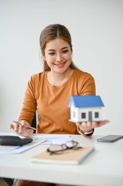 Asyalı güzel bir emlakçının portresi. Masasında bir ev modelini tutuyor ve ona bakıyor..