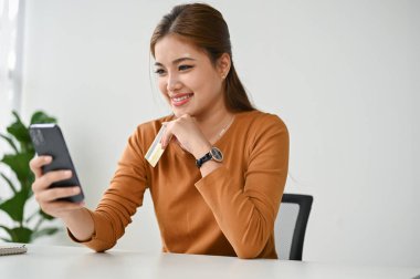 Güzel ve mutlu bir Asyalı kadın kredi kartını akıllı telefonuyla alışveriş uygulamasına bağlıyor, evden online alışveriş yapmaktan hoşlanıyor..