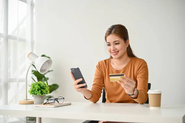 一位快乐的亚洲女人坐在桌旁 把她的信用卡和一个购物应用软件连接起来 — 图库照片