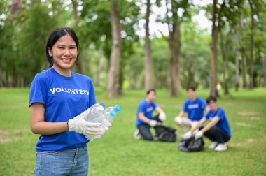 Üniformalı, güzel ve mutlu bir Asyalı kadın gönüllü halka açık bir parkta plastik şişelerle duruyor ve ekibiyle birlikte bir parkı temizlemek için gönüllü olarak çalışıyor..