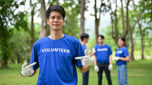 一位快乐而英俊的身穿制服的年轻亚洲男性志愿者站在公园里 与他的朋友们一起参与清理公园的志愿工作 — 图库照片