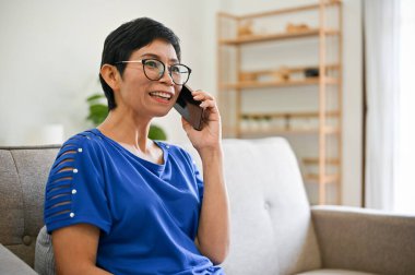 Mutlu, orta yaşlı, Asyalı bir kadın oturma odasındaki kanepede dinlenirken torunuyla telefonda konuşuyor..