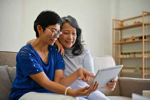 stock image Two happy and lovely retired Asian female friends are watching online clips on social media on a tablet while relaxing on a sofa in the living room together.