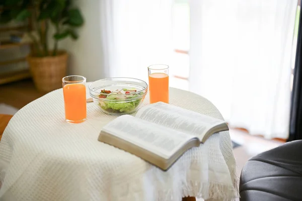 Close-up image of an opened book, a salad bowl, and two glasses of orange juice are on a table in a minimalist dining room.