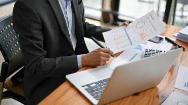 Resmi bir iş elbisesi içindeki akıllı ve profesyonel bir işadamının ya da erkek analistin yakın plan görüntüsü masasındaki mali raporları inceleyip analiz etmektedir..