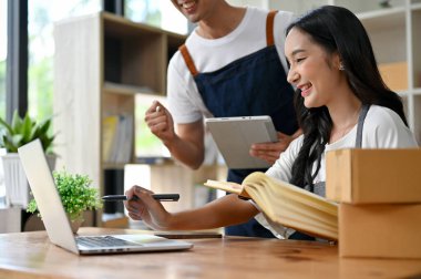 Güzel bir Asyalı genç kadın çevrimiçi satıcının yakın plan resmi dizüstü bilgisayarına bakıyor, siparişleri kontrol ediyor ve alışveriş sitesindeki yeni ürünleri güncelliyor, ve erkek arkadaşıyla çalışıyor..