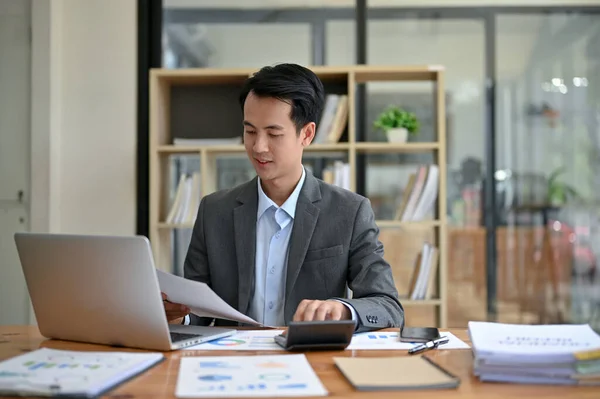 stock image A professional and handsome millennial Asian businessman focuses on business financial reports, working at his desk in the office.