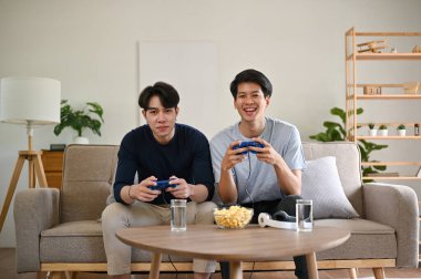 İki neşeli ve mutlu Asyalı genç erkek arkadaş oturma odasındaki bir kanepede video oyunu oynuyorlar. Oyun kumandası, oyun kumandası