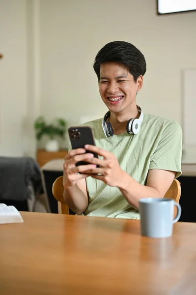 一位穿着休闲装的快乐的亚洲年轻人 喜欢坐在餐桌旁 通过智能手机与朋友们聊天 — 图库照片