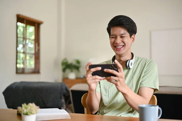 一位穿着休闲装的快乐的亚洲年轻人 喜欢坐在客厅的桌子旁 用智能手机玩手机游戏 — 图库照片