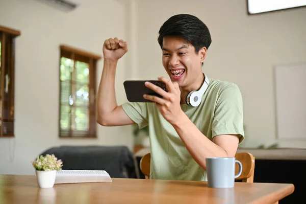 一位身穿舒适衣服的兴奋而喜庆的亚洲年轻人举起手 一边庆祝一场体育比赛的胜利 一边在客厅的桌子旁玩手机游戏 — 图库照片