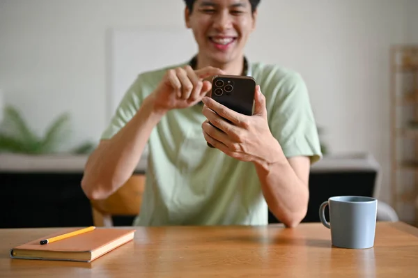 一位快乐的亚洲年轻人正坐在客厅的桌子旁 一边用智能手机 在社交媒体上滚动 与朋友聊天 一边在应用程序上享受网上购物的乐趣 — 图库照片