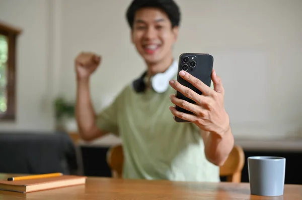 一位兴奋的亚洲年轻人举起手来 一边庆祝一场体育比赛的胜利 一边在客厅里的桌子旁 一边用手机玩手机游戏 — 图库照片