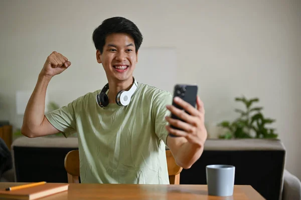一名兴奋的亚洲年轻人举手庆祝一场体育比赛的胜利 或在客厅里的桌子旁 用手机玩手机游戏 — 图库照片