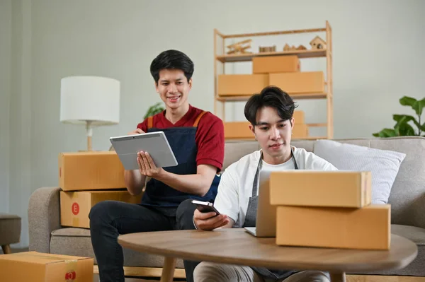 A happy young Asian gay couple, online shop owners, updating new products on the website, preparing shipping orders, and working at home together. online business, e-commerce, LGBT business partners
