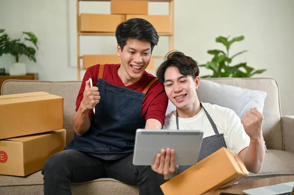 A cheerful young Asian gay couple checks their online product orders on a tablet and celebrates their orders success while working at home together. LGBT couple, e-commerce, online shop, online seller