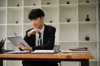 Resmi bir iş elbisesi içinde akıllı ve yakışıklı bir Asyalı işadamı ofisinde masasında otururken tabletinden bir şeyler okumaya odaklanır..